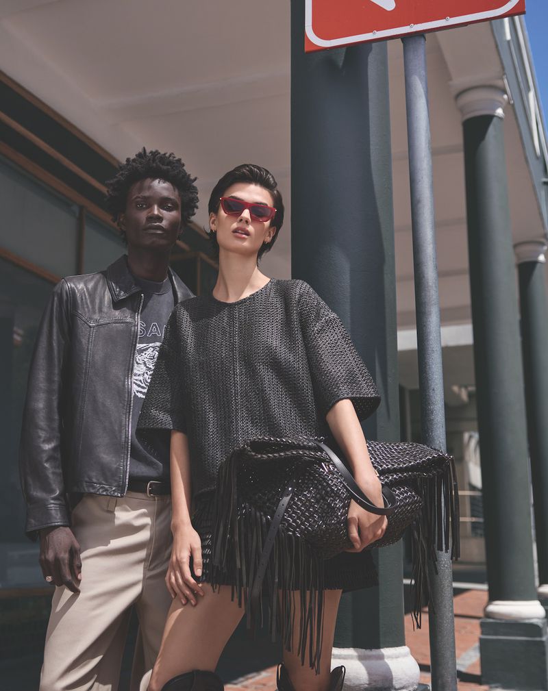 Two fashion models pose in front of a building, wearing stylish clothing and sunglasses, with a large fringed handbag as an accessory.