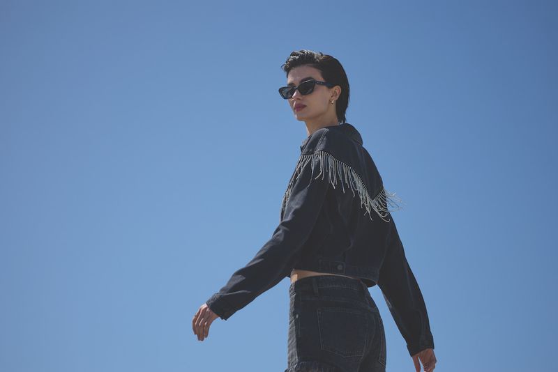 Woman in a black denim jacket with silver fringe and sunglasses, against a blue sky. Low angle, fashion photography.