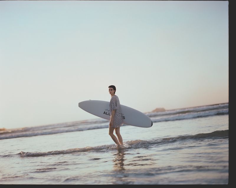 Adult woman in a floral dress walks out of ocean carrying a surfboard at sunset. Coastal scenery, water reflections.