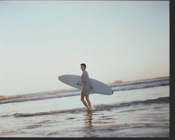 Adult woman in a floral dress walks out of ocean carrying a surfboard at sunset. Coastal scenery, water reflections.