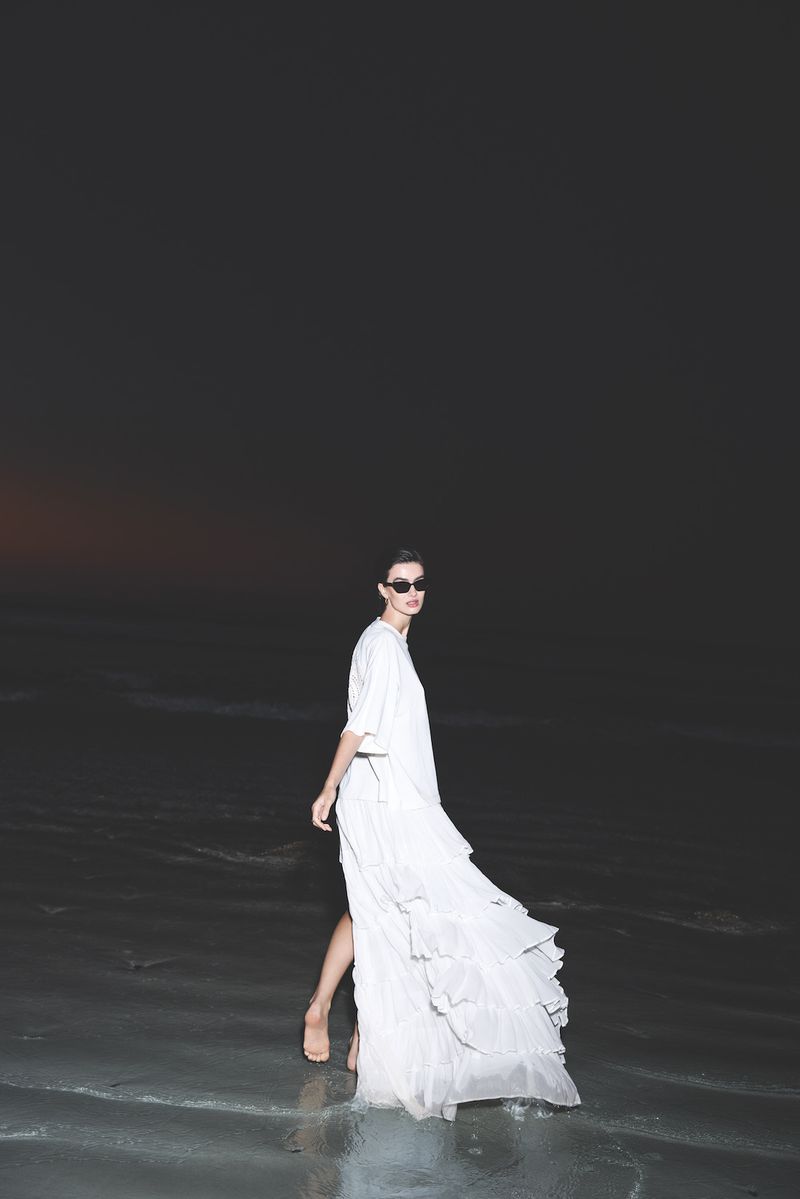 Fashion shot of an adult woman wearing a long, flowing white dress and sunglasses walking on a reflective, wet surface in a dark environment.