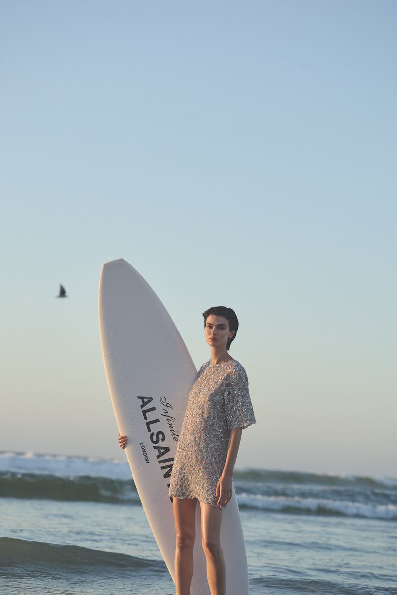 A woman stands on a beach holding a surfboard, wearing a short, shimmering dress, with waves and the sky in the background.