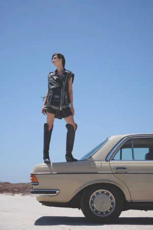 A woman in a vest and boots poses atop a vintage car in an outdoor setting with a clear sky; a fashion-editorial image.