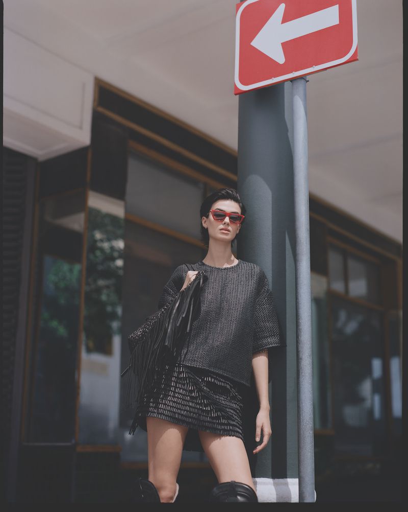 Fashionable woman in tweed mini skirt and top stands in front of street sign, carrying jacket. She wears sunglasses and high boots.