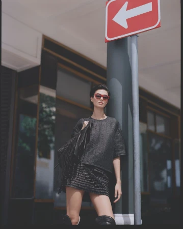 Fashionable woman in tweed mini skirt and top stands in front of street sign, carrying jacket. She wears sunglasses and high boots.