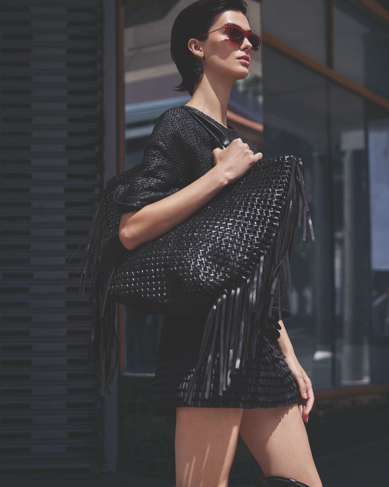 Elegant woman in black dress and red sunglasses carrying a large woven fringe handbag on a city street.