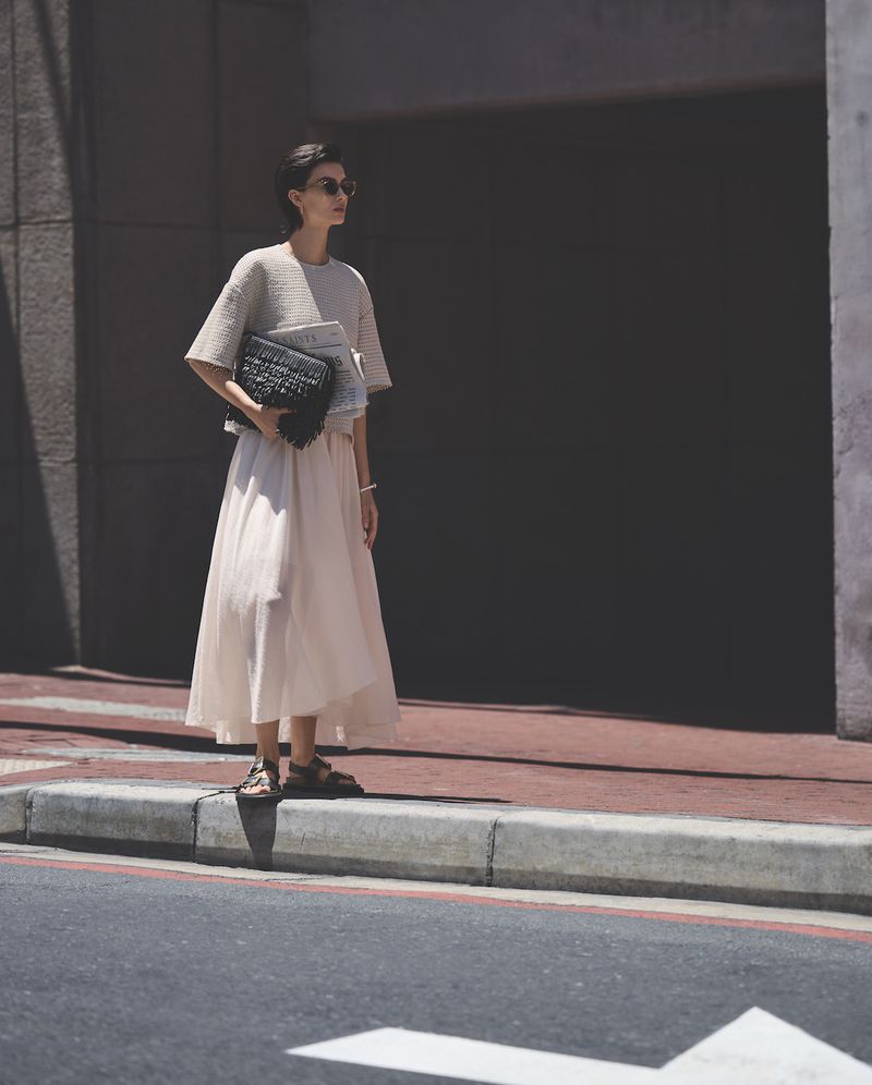 A woman in a cream-colored skirt and top stands on a city street holding a black purse and magazine, bathed in bright sunlight.