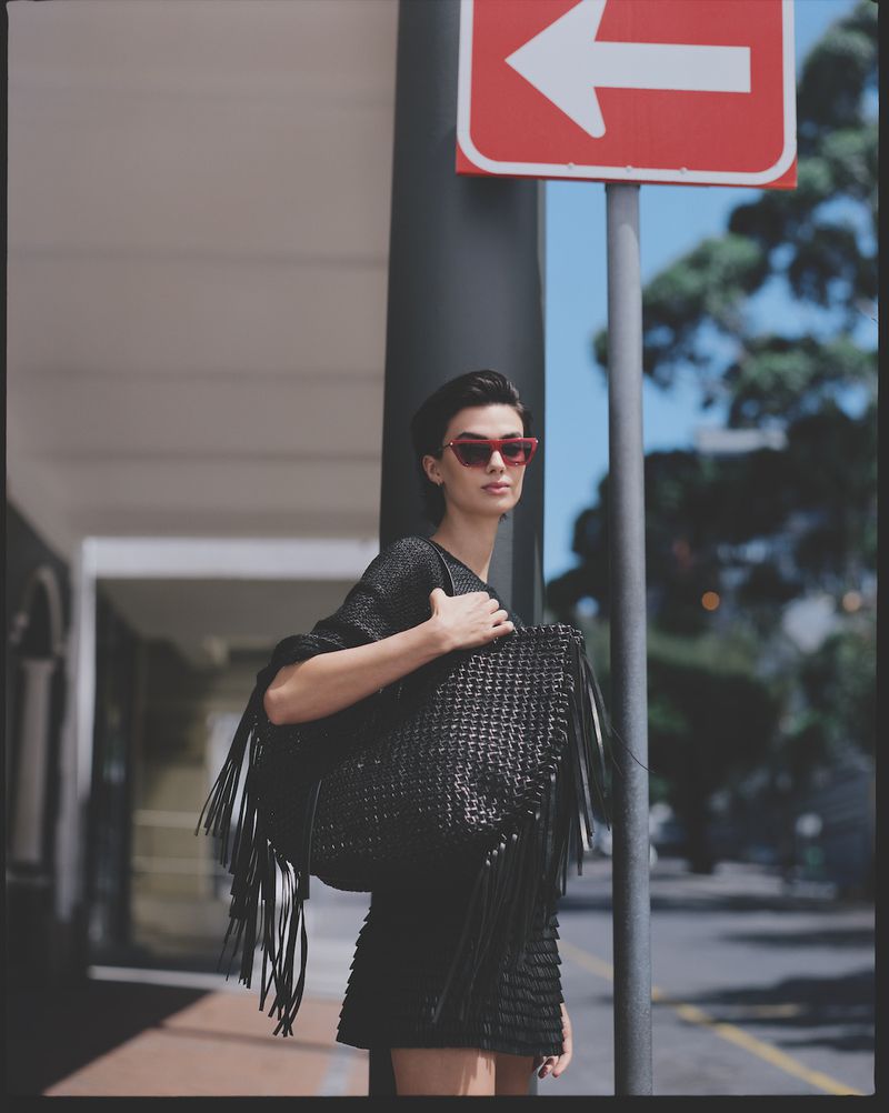 Stylish adult woman in a short black dress and red sunglasses carrying a black woven fringe bag on an urban street with red sign.