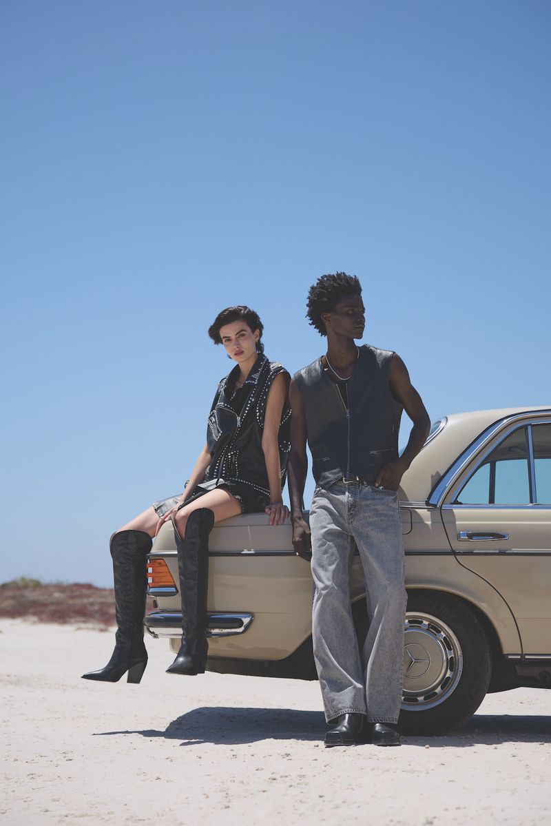 Two fashion models pose on a vintage tan car against a clear blue sky, wearing stylish black vests, jeans, and thigh-high boots.