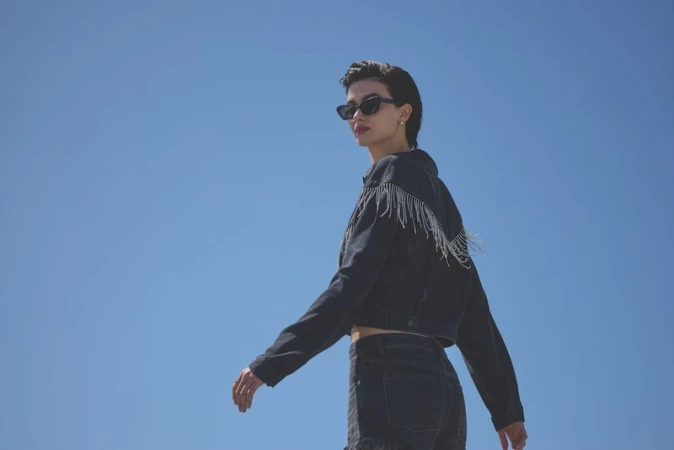 A model wearing dark sunglasses and a black denim jacket with fringe details against a clear blue sky background.