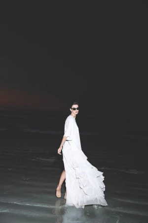 Model wearing a white dress and sunglasses walking on a sandy beach at night with ocean waves in the background.