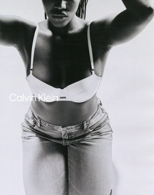 Monochrome studio shot of an adult female fashion model wearing a white bra and light denim skirt, posing against a white backdrop.