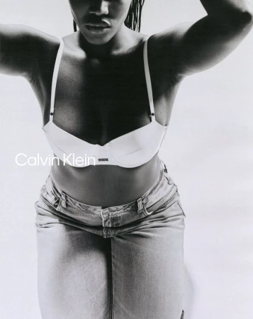 Monochrome studio shot of an adult female fashion model wearing a white bra and light denim skirt, posing against a white backdrop.