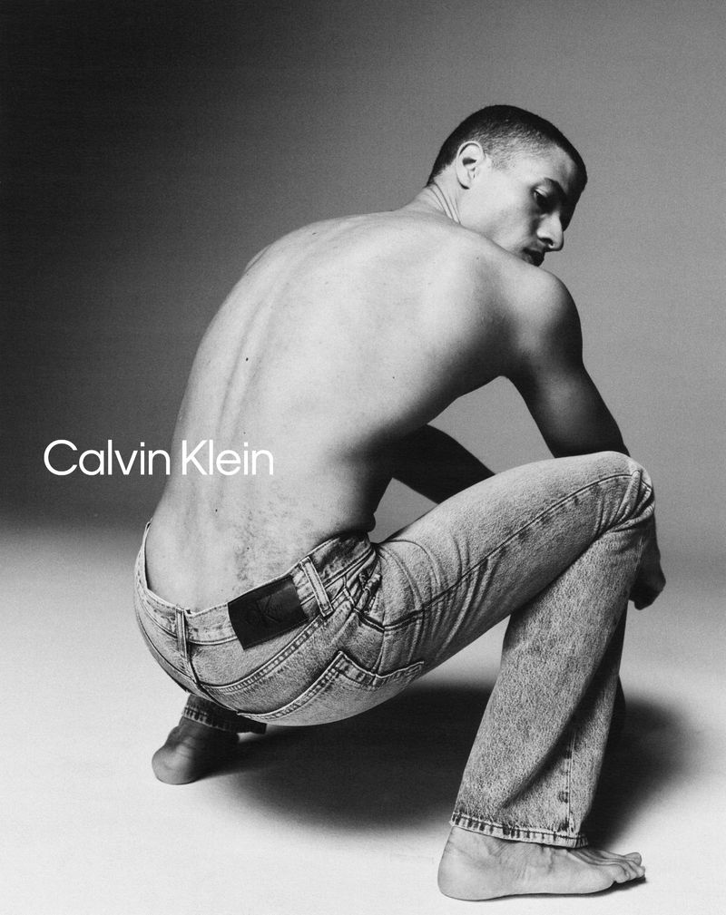 Black-and-white Calvin Klein ad featuring a shirtless man in jeans crouching while looking over his shoulder.