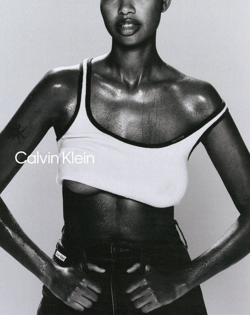 A Black woman in a white tank top and black bottoms poses against a grey backdrop, showcasing a Calvin Klein design.