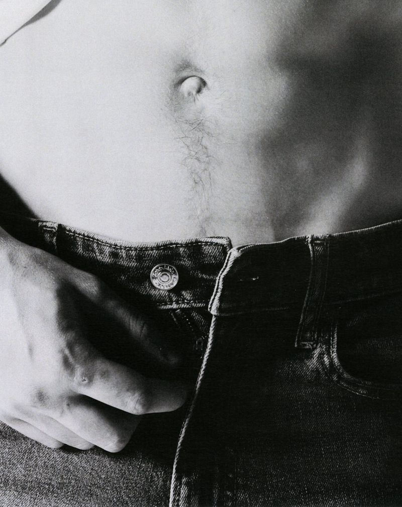 Close-up shot of a person's torso and hand, wearing jeans, in black and white. Focus on the waist and hand adjusting the waistband.
