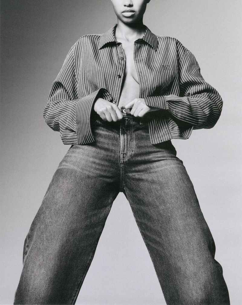 Model wearing a striped button-down shirt and relaxed denim jeans against a solid light grey studio backdrop.