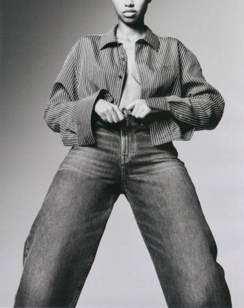 Model wearing a striped button-down shirt and relaxed denim jeans against a solid light grey studio backdrop.