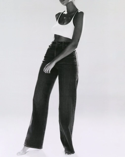 An African-American woman is posing in a photo studio wearing a white tank top and black jeans against a bright white background.