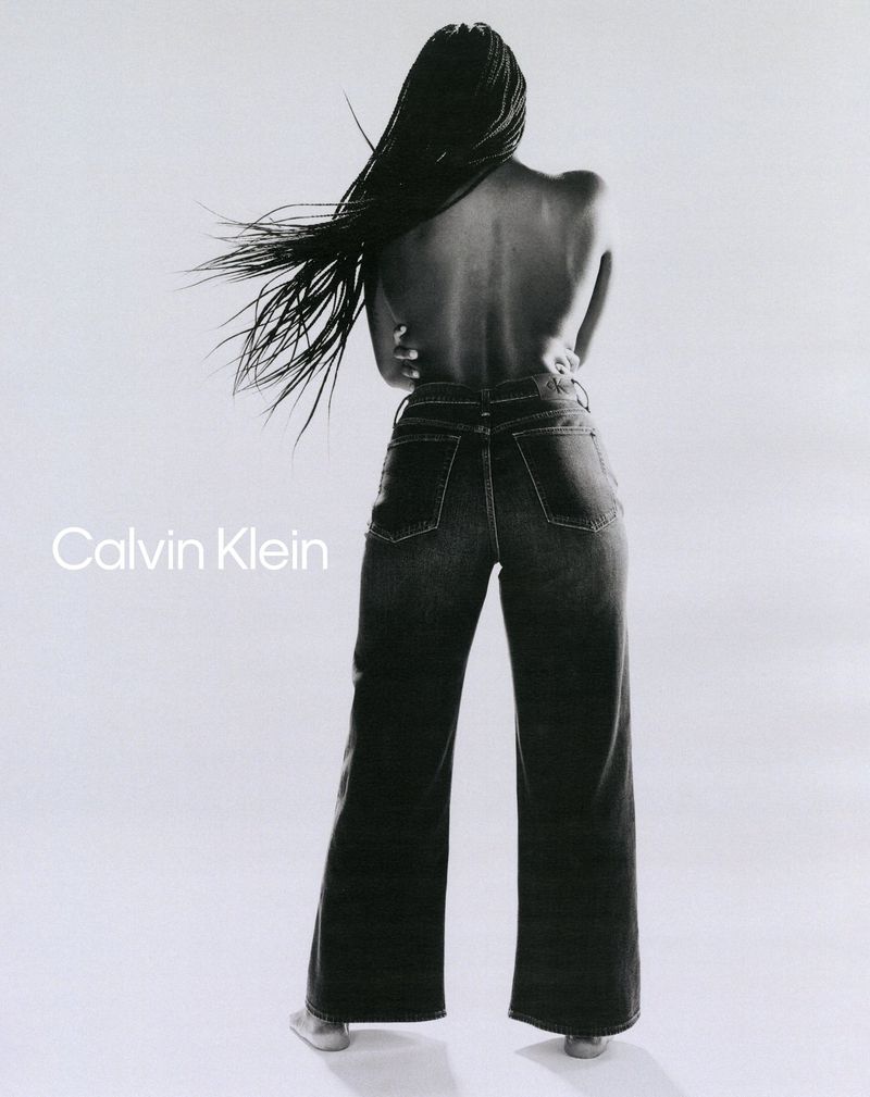 Model with long braided hair wearing dark denim jeans posing against a white background for Calvin Klein.