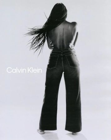 Model with long braided hair wearing dark denim jeans posing against a white background for Calvin Klein.