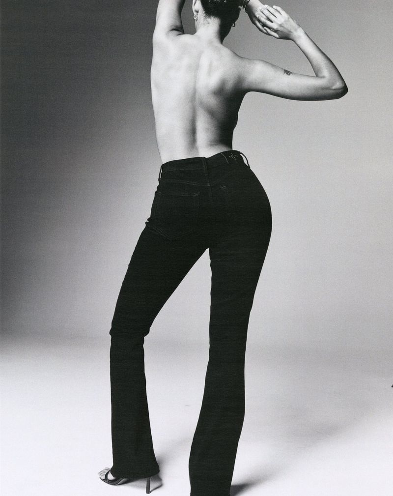 Back view of an adult female model wearing black denim jeans and high heels, posing in a studio with her arms raised.