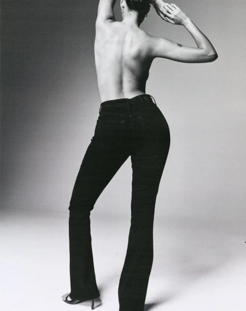 Back view of an adult female model wearing black denim jeans and high heels, posing in a studio with her arms raised.