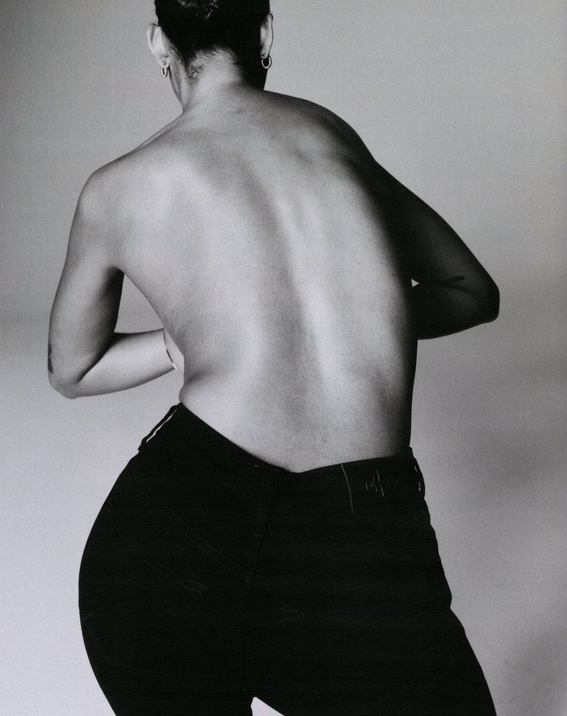 Black and white studio shot of a slender adult female posing with bare back to the camera, wearing black jeans against a plain backdrop.