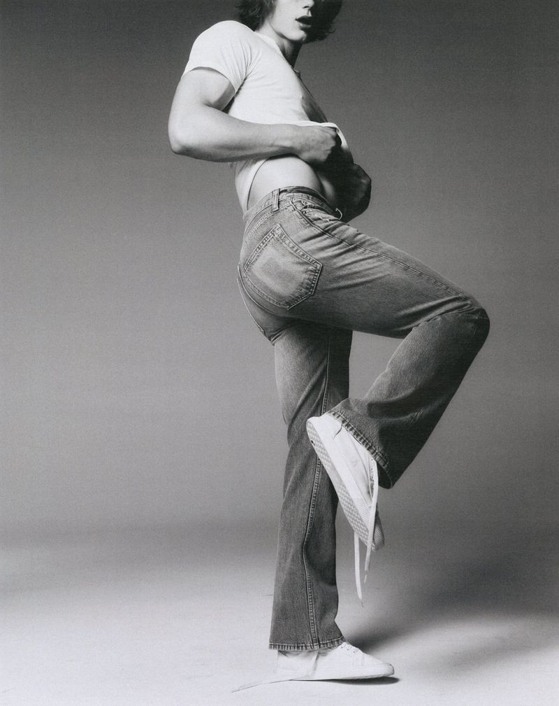 An adult male poses in jeans and a T-shirt, lifting his leg and shirt in a black and white studio fashion shot against a grey backdrop.