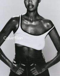 Model wearing a white tank top and dark denim jeans posing against a neutral grey studio background with Calvin Klein branding.