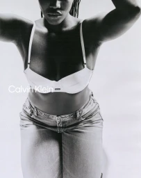 Model wearing a white bra and denim jeans against a plain white seamless studio background for Calvin Klein.