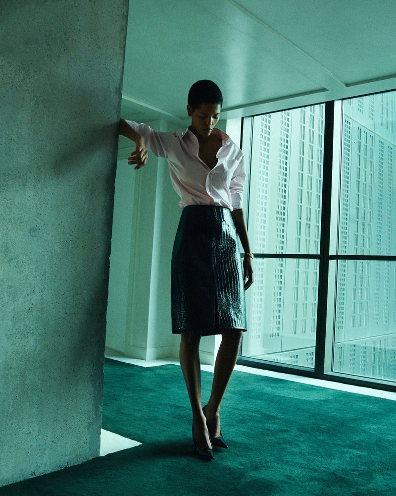 Woman in pink shirt and dark skirt stands near window and wall inside a building with green carpet and view of buildings outside.