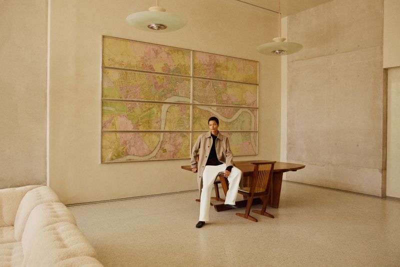 Stylish woman poses indoors near a table and art, bathed in soft, neutral lighting, with a couch subtly placed on the left.