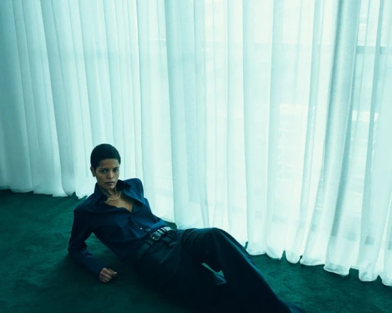 A woman with short hair poses on a green carpet in a navy blue button-down shirt and trousers, illuminated by natural light.