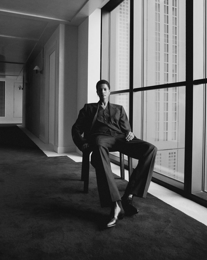 Black and white portrait of a woman in a suit sitting indoors by a large window overlooking a city. Modern, high-fashion.