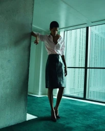 Model standing on a green carpet in a room with large windows against a city building exterior, wearing a pink shirt and dark leather skirt.