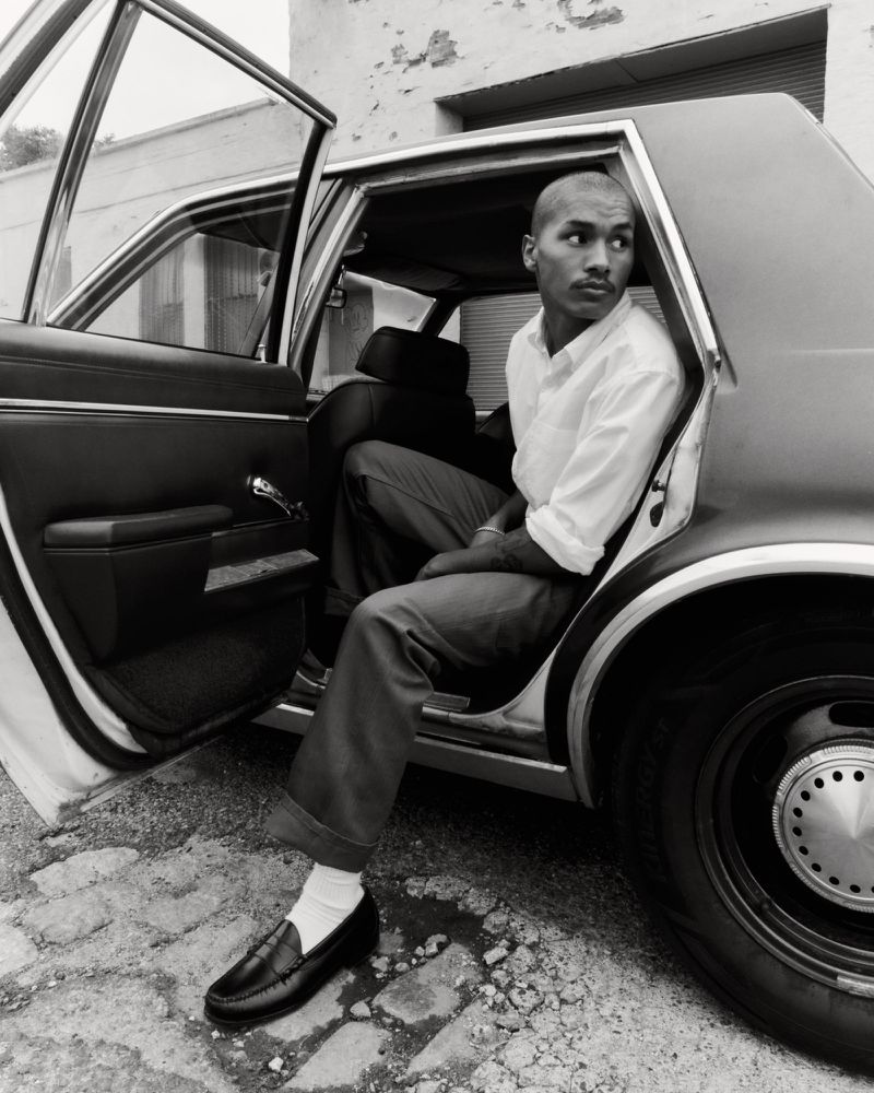 Monochrome image of a stylish adult male sitting in the open back seat of a vintage car, wearing loafers and a button-down shirt.