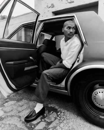 Monochrome image of a stylish adult male sitting in the open back seat of a vintage car, wearing loafers and a button-down shirt.