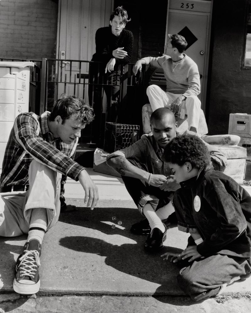 Monochrome lifestyle editorial showing a group of men gathered outdoors playing dice on a sidewalk