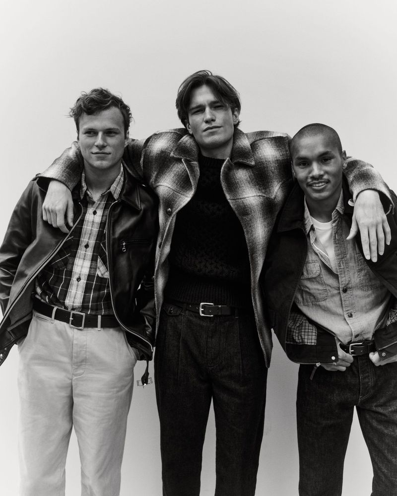 Three stylish adult men pose indoors wearing jackets, button-down shirts, and trousers in a medium studio shot. 