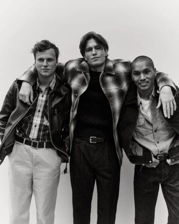 Three stylish adult men pose indoors wearing jackets, button-down shirts, and trousers in a medium studio shot. 