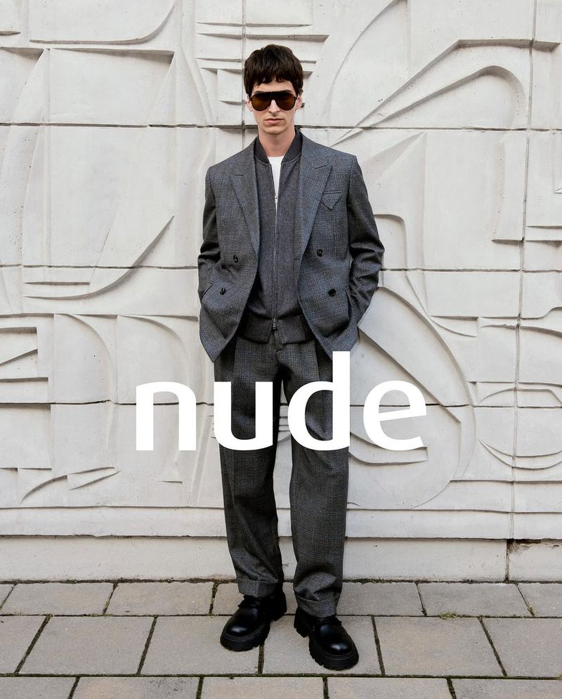 A man in a grey suit and sunglasses poses against a patterned wall and stone floor. He is looking directly at the camera.