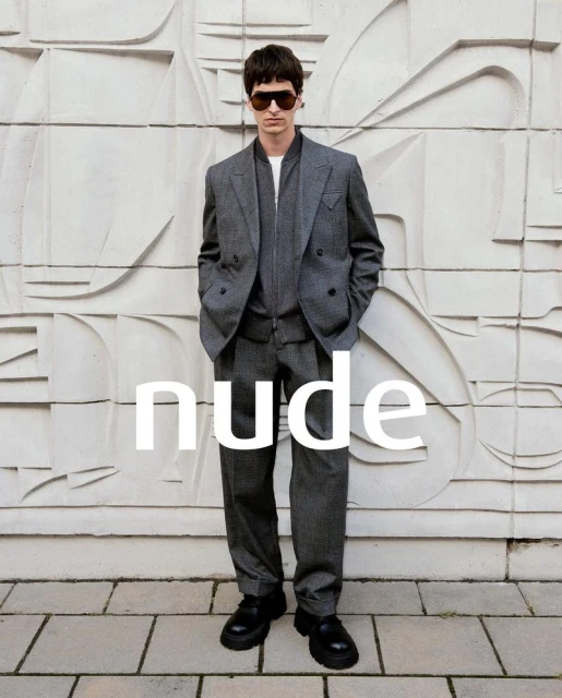 A man in a grey suit and sunglasses poses against a patterned wall and stone floor. He is looking directly at the camera.