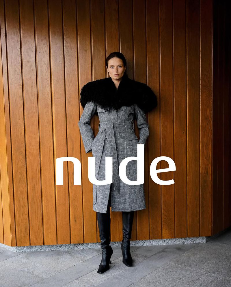 Woman in gray trench coat with black fur collar and black knee-high boots posing in front of a wooden paneled wall indoors.
