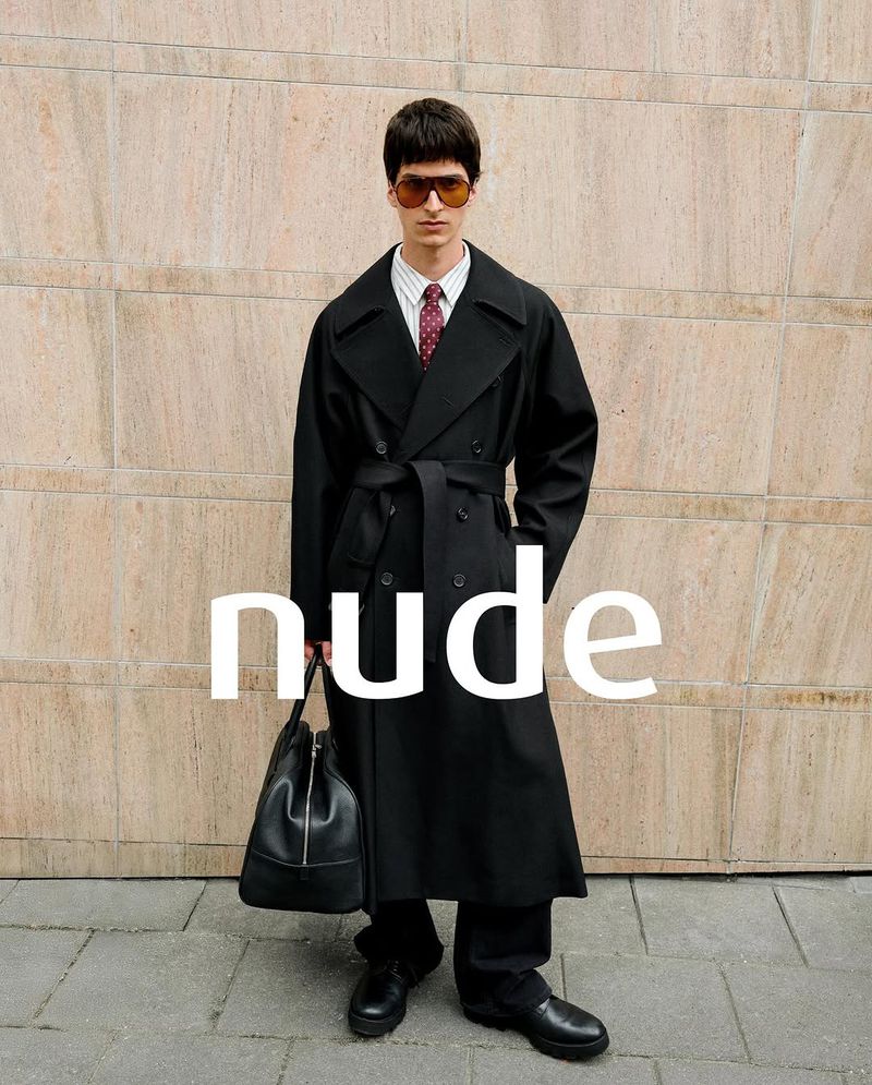 Man in a black trench coat with a bag, standing against a textured wall in an outdoor urban setting, showcasing a fashion-editorial style.