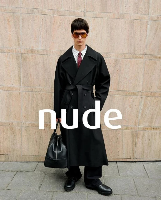 Man in a black trench coat with a bag, standing against a textured wall in an outdoor urban setting, showcasing a fashion-editorial style.