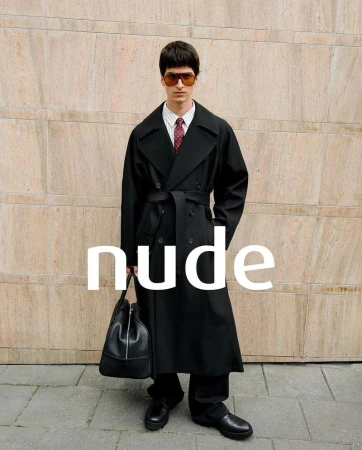 Man in a black trench coat with a bag, standing against a textured wall in an outdoor urban setting, showcasing a fashion-editorial style.