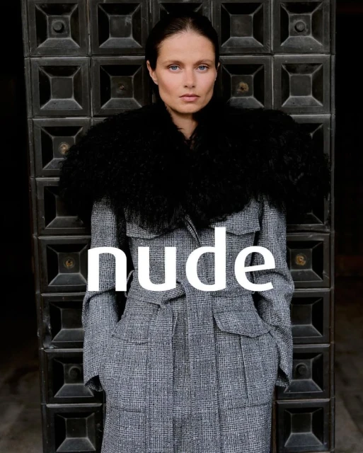 A woman in a gray patterned coat with a black fur collar poses against a dark, geometrically patterned door, looking directly at the viewer.