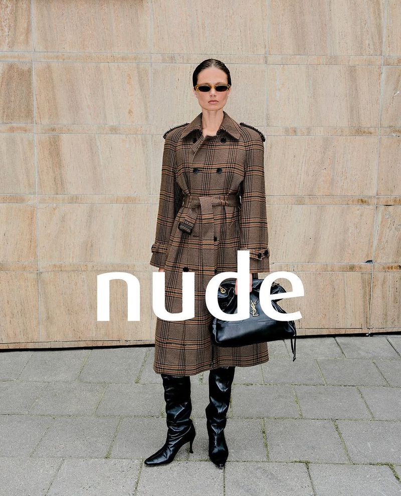Model wearing a brown plaid belted trench coat, black leather boots, and sunglasses standing against a stone wall with text overlay.