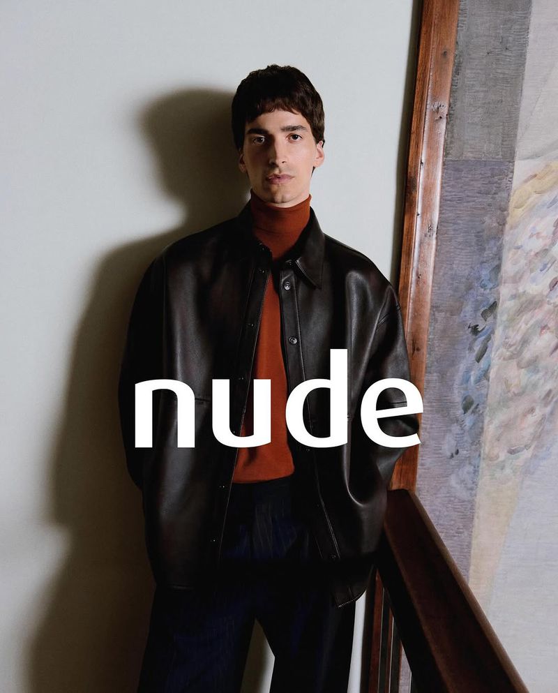 Portrait of a brunette man in a dark leather jacket and orange turtleneck, standing against a neutral background with a painting.